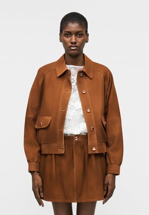 Mujer con una chaqueta marrón abotonada y falda a juego, con una blusa blanca de encaje floral debajo, posando contra un fondo liso.