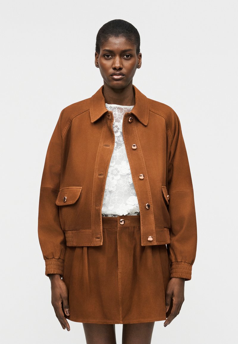 Γυναίκα που φοράει καφέ σακάκι με κουμπιά και ταιριαστή φούστα, με λευκό δαντελωτό τοπ με floral λεπτομέρειες από κάτω, στέκεται μπροστά σε απλό φόντο.