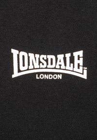 Crna tkanina s bijelim natpisom "LONSDALE LONDON". Slova imaju hrabar, zakrivljen dizajn, postavljena na teksturiranu pozadinu.