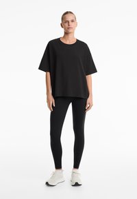 Fekete, oversized póló puha anyagból, fekete leggings-szel párosítva. A modellen világos színű sportcipő van. Minimalista dizájn, minta nélkül.