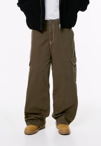 Pantaloni cargo marroni larghi con tasche laterali, indossati con stivali beige, una camicia bianca e un giubbotto nero con le mani nelle tasche.