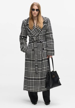 Mujer con abrigo de pata de gallo en blanco y negro con cinturón, pantalones negros, gafas de sol y llevando un bolso negro con detalles dorados.