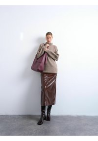 Femme appuyée contre un mur blanc, portant un pull beige, une jupe midi marron brillante, des bottes noires, tenant un grand sac à main bordeaux sur l'épaule.