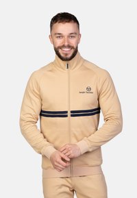 Beige zip-jakke med høj krave, med marinebl�å vandrette striber og logo på venstre bryst. Blødt stof, atletisk pasform, ribbede manchetter.