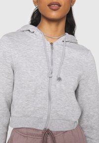 Sudadera gris con cremallera, capucha ajustable, diseño corto, textura suave y cremallera en contraste. Llevada sobre pantalones rosa claro.