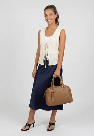 Femme portant un gilet en maille sans manches crème et une jupe midi bleu marine, tenant un sac à main en cuir beige et portant des sandales noires à talons, souriante.