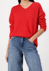 Suéter rojo acanalado con cuello en V y mangas arremangadas, combinado con jeans de mezclilla azul de cintura alta. Punto texturizado y ajuste relajado.