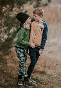 Dwoje dzieci stoi blisko siebie. Jedno ma na sobie zieloną bluzę z kapturem, spodnie w wzory oraz czarną czapkę beanie. Drugie nosi beżowo-niebieską kurtkę i ciemne spodnie.