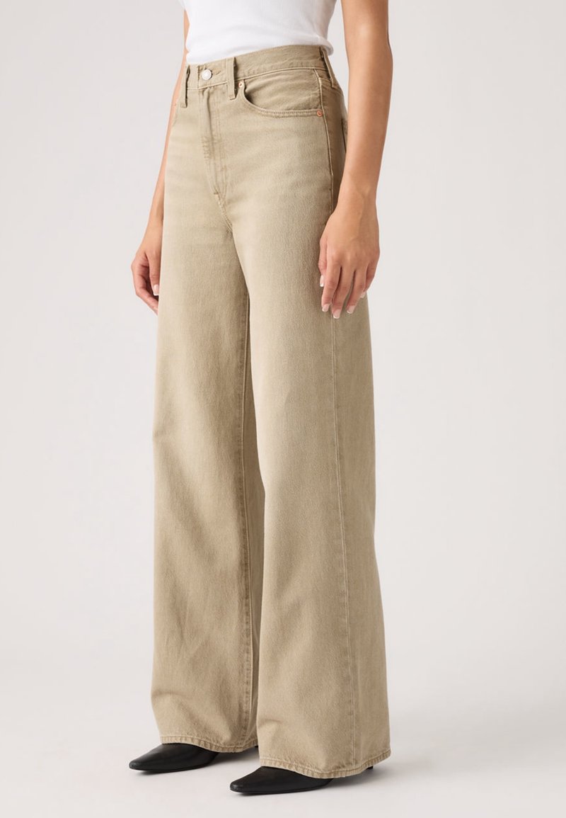 Jean beige taille haute à jambes larges porté avec des chaussures noires pointues et un haut blanc rentré, sur un fond uni.