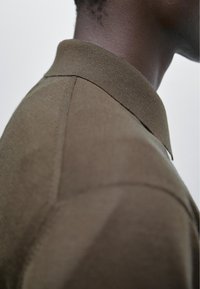 Short-sleeved, olive green knit polo shirt with a classic collar, featuring smooth texture and detailed shoulder seams.