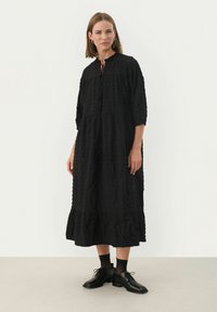 Schwarzes, langärmeliges Kleid aus strukturiertem Stoff mit gerafftem Ausschnitt und ausgestelltem Saum. Tragen mit schwarzen Schnürschuhen und Knöchelsocken.