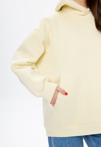 Helle gelbe Kapuzenjacke mit einer Fronttasche, gerippten Bündchen und einer Zugkordel an der Kapuze. Weicher Stoff, lässige Passform, detaillierte Nähte sichtbar.