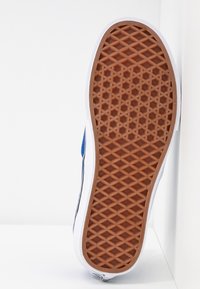 Semelle d'une basket avec un motif de crampons en forme de gaufre marron, une semelle intermédiaire blanche et une tige bleue visible sur le côté.