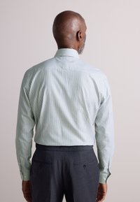 Pruhovaná košile světle zelené a bílé barvy s klasickým límcem a dlouhými rukávy, doplněná tmavě šedými kalhotami s knoflíkovými detaily.