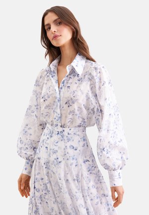 Vrouw die een witte blouse en rok draagt met lichtblauwe bloemmotieven en volumineuze lange mouwen, staand tegen een effen achtergrond.