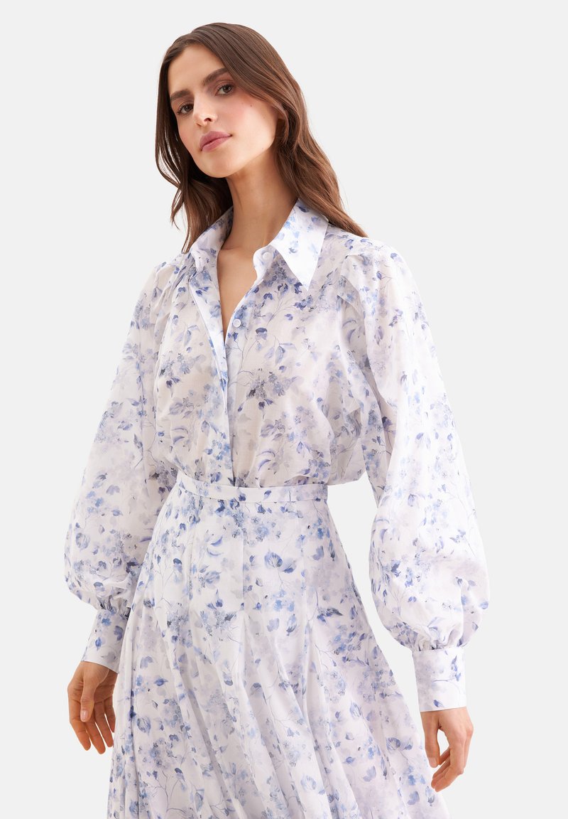 Femme portant un chemisier blanc et une jupe avec des motifs floraux bleu clair et des manches longues volumineuses, debout devant un fond uni.