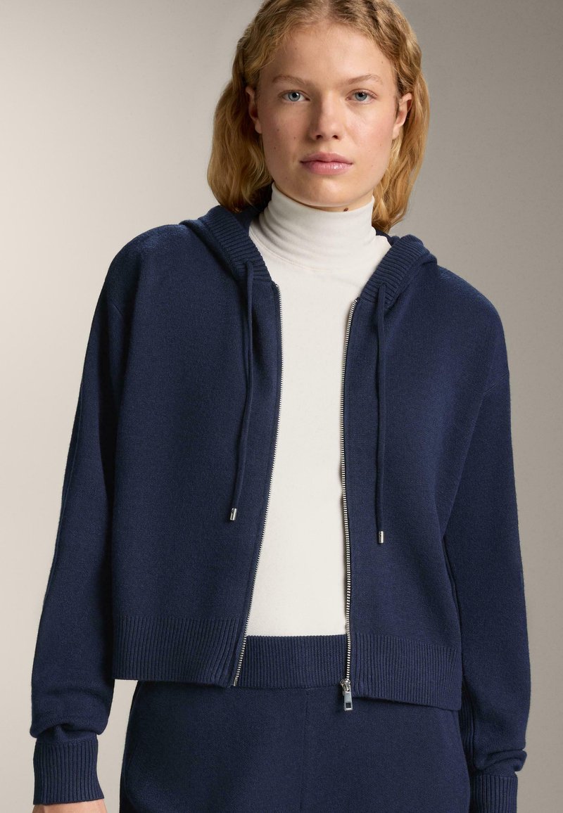 Námornícky modrá bunda na zips s kapucňou, rebrovanými manžetami a pásom. Mäkka textúra s kontrastným bielym rolákom pod ňou.