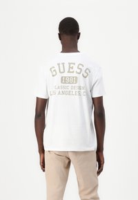 Hvit bomull t-skjorte med "GUESS 1981" og "CLASSIC DESIGN LOS ANGELES, C." trykt i beige på baksiden. Korte ermer, rund hals.