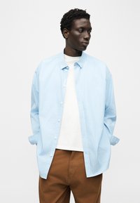 Νέος άντρας με κοντά σγουρά μαλλιά φορώντας ένα ανοιχτό μπλε ριγέ πουκάμισο πάνω από λευκό t-shirt και καφέ παντελόνι, με τα χέρια στις τσέπες, κοιτάζοντας δεξιά.