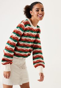 Strickpullover in rot, grün und cremefarbenem Wellenmuster mit einem gerippten Ausschnitt und Bündchen, kombiniert mit einem cremefarbenen Cordrock mit Knöpfen.