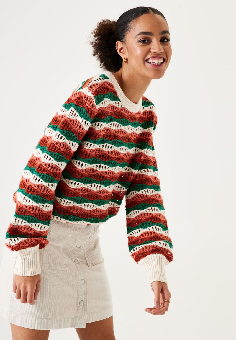 Strickpullover in rot, grün und cremefarbenem Wellenmuster mit einem gerippten Ausschnitt und Bündchen, kombiniert mit einem cremefarbenen Cordrock mit Knöpfen.