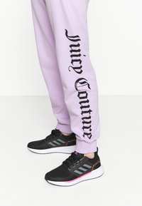 Černé sportovní boty s texturovaným síťovaným svrškem, šedými akcenty a růžovými detaily, doplněné světle fialovými tepláky s černým logem.