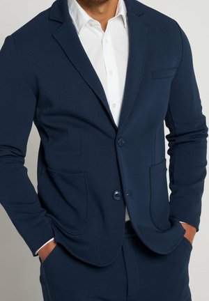 Mann trägt ein tailliertes marineblaues Sakko mit zwei Knöpfen und aufgesetzten Taschen über einem weißen Hemd, Hände in den Taschen.