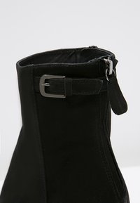 Bottine en daim noir avec un bout pointu, fermeture éclair et un détail boucle métallique rectangulaire sur le côté. Texture lisse.
