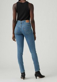 Jeans ajustados de color azul con una cintura de altura media, que presentan un parche de cuero en la parte trasera, cinco bolsillos y detalles de costura sutiles, combinados con una blusa sin mangas negra.