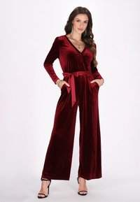 Burgundy bársonyfelső overál, hosszú ujjakkal, V-kivágással, széles lábakkal, derékmegkötővel és oldalsó zsebekkel. Simább textúra és elegáns megjelenés.