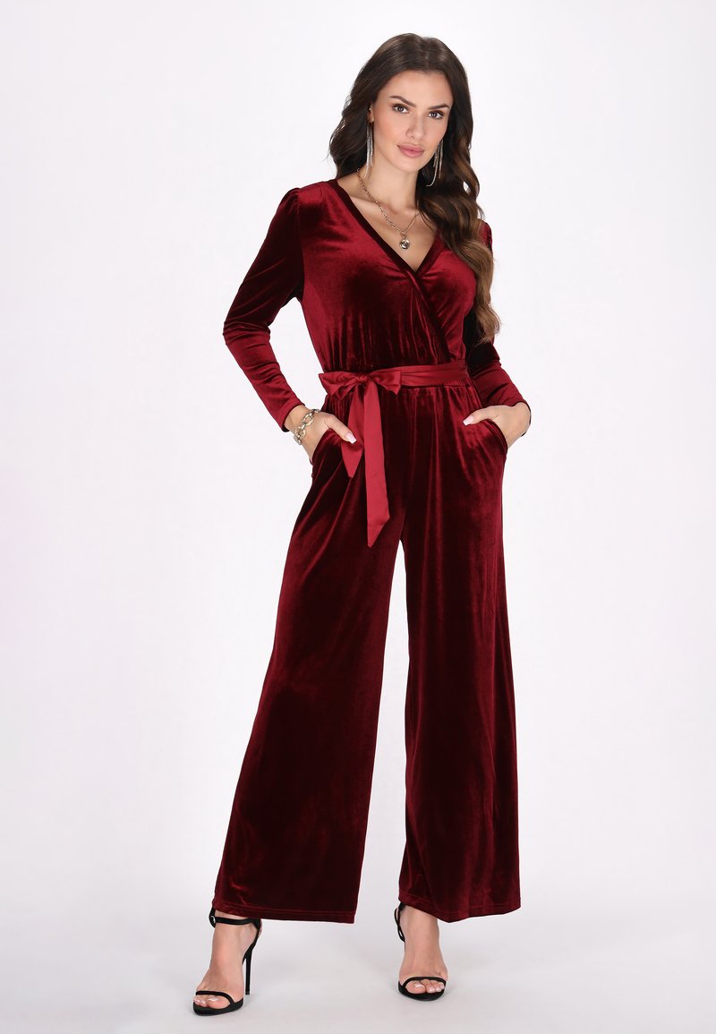 Burgundyröd sammet jumpsuit med långa ärmar, V-ringning, vida byxor, midjeband och sidofickor. Slät textur och elegant finish.