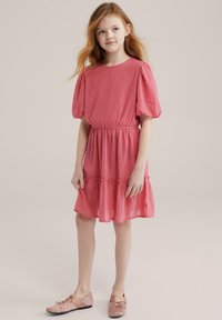 Vestido corto rosa con mangas abullonadas y cintura elástica. Presenta un dobladillo en capas y una tela texturizada. Combinado con zapatos planos de un rosa claro.