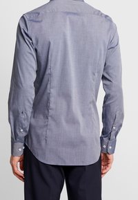 Mężczyzna w jasnoniebieskiej koszuli z długim rękawem i ciemnych spodniach, pokazany od tyłu na jednolitym białym tle.