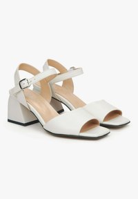 Witte leren haksandalen met een vierkante neus, verstelbare enkelband en een chunky hak. De buitenzool heeft een contrasterende zwarte afwerking.