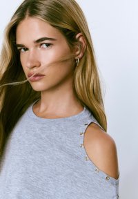 Top gris de hombros al aire con acentos de hardware dorado y una textura suave. Presenta un escote relajado y cortes en los hombros. Tela suave.