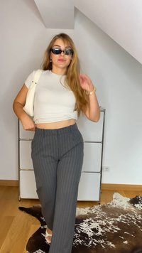 T-shirt bianca cropped, pantaloni grigi a righe, occhiali da sole neri e una borsa bianca. Pavimento in legno e tappeto decorato sullo sfondo.