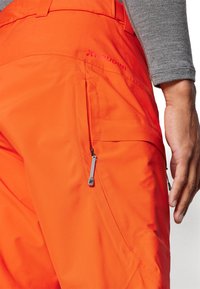 Orange Freizeit-Hosen aus strapazierfähigem Material, mit einer Reißverschlusstasche, glatter Textur und kontrastierendem Logo-Detail am Bund.