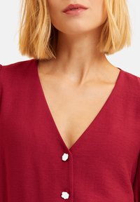 Burgunderfarbene Bluse mit tiefem V-Ausschnitt, verziert mit weißen Blumenknöpfen und weichem Textilgewebe. Sichtbare Hautfarbe im Ausschnittbereich.