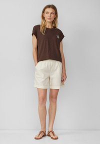 Camiseta marrón de manga corta con un corte relajado, combinada con pantalones cortos color crema con dobladillo enrollado y sandalias planas marrones. Diseño simple y tela suave.