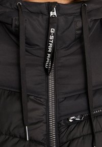 Svart vadderad jacka med dragkedjestängning, texturerat tyg, justerbara dragsko och en sidoficka med dragkedja. Har varumärkets logotyp på dragkedjan.