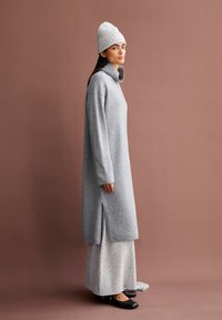 Lichtgrijze lange sweaterdress met hoge kraag, zijsplitten en ribbelstructuur, gecombineerd met een crèmekleurige gebreide beanie en zwarte schoenen.
