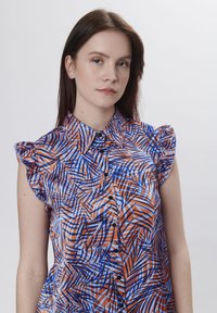 Gekraagde blouse met gerimpelde mouwen, voorzien van een kleurrijk patroon van blauwe en oranje geometrische vormen, bevestigd met zwarte knopen.