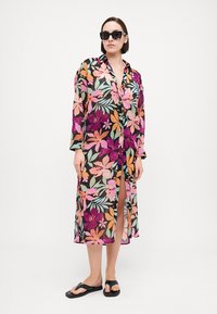 Kimono noir transparent avec un imprimé floral coloré. Manches longues, fermeture à boutons et fente jusqu'à la cuisse. Porté avec des tongs noires et des lunettes de soleil.