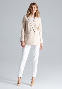 Beige op maat gemaakte blazer met dubbele knopen, gecombineerd met witte getailleerde broeken en naakthele hoge hakken, op een lichtgrijze achtergrond.