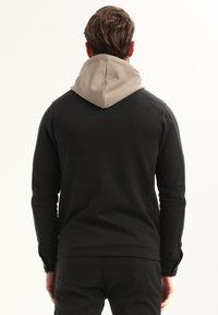 Uomo che indossa un sudario nero con cappuccio beige, in piedi con le spalle alla telecamera su uno sfondo bianco semplice.