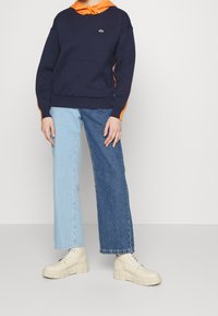 Person wearing a navy hoodie with orange hood, two-tone blue jeans, and cream platform lace-up boots, standing against a plain background.