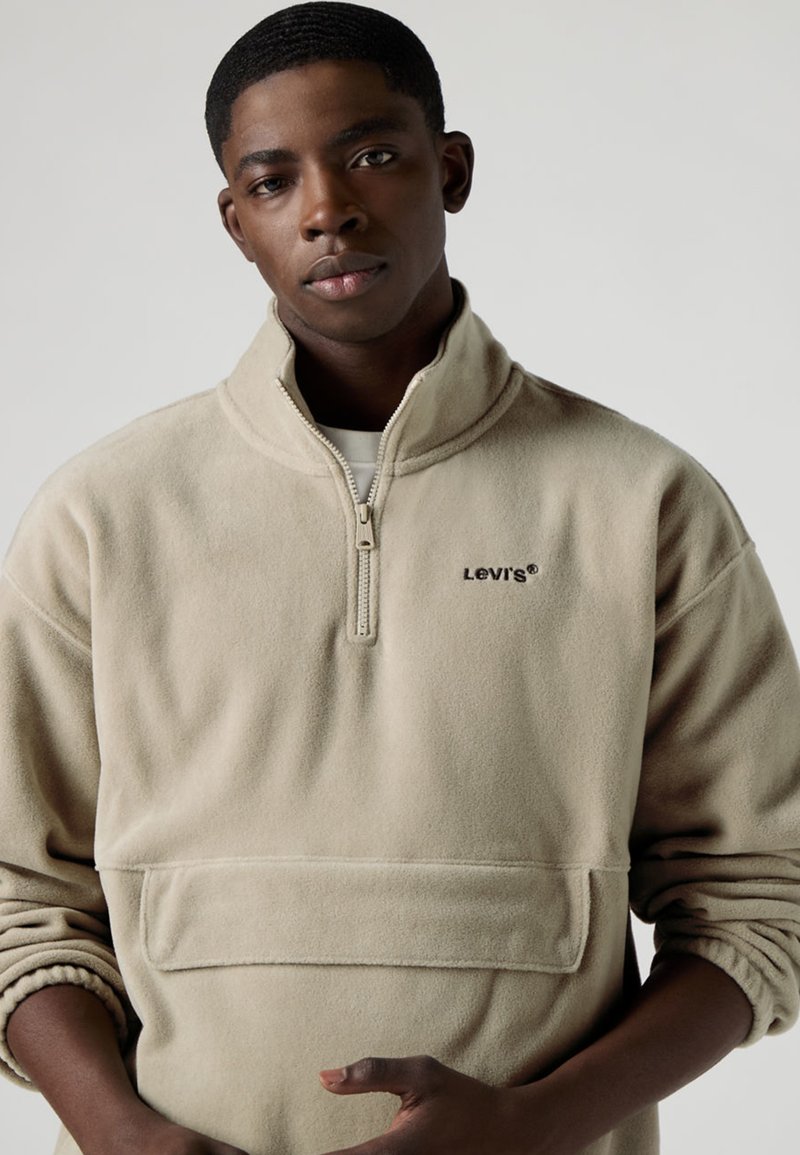 Beige fleecetröja med kvartszipp, rundad krage, framficka och broderad Levi's-logotyp i mörk färg. Mjuk textur och avslappnad passform.