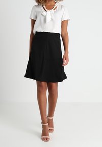 Blusa bianca con un dettaglio al collo a fiocco abbinata a una gonna nera a campana. Il modello indossa tacchi nudi, mostrando un tessuto liscio e un design strutturato.