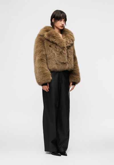 Γυναίκα που στέκεται, φορώντας καφέ χνουδωτό γούνινο μπουφάν, μαύρο παντελόνι με φαρδιά πόδια και μαύρα μυτερά παπούτσια, μπροστά σε απλό λευκό φόντο.