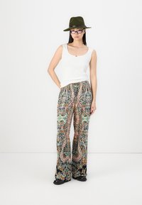 Top sin mangas blanco, pantalones de pierna ancha con diseño de paisley en verde, naranja y azul, zapatos negros y un sombrero verde olivo.